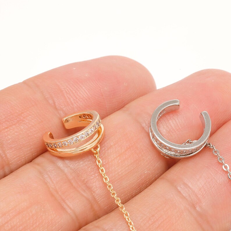 May include: Two ear cuffs with attached chains, one in a rose gold tone and one in a silver tone. Each cuff features a double-band design with a row of small, clear, sparkling stones embedded along the outer band, connected to a thin metal chain.