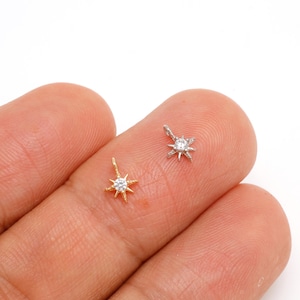 May include: Two small starburst pendants, one gold-toned and one silver-toned, each featuring a central clear gemstone. The pendants are displayed against a neutral background, showcasing their delicate design and sparkle. These are ideal for jewelry making.