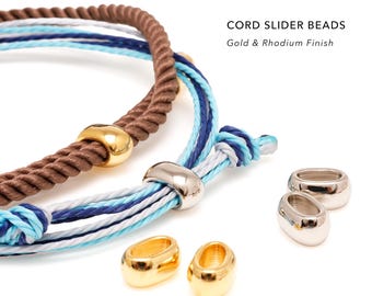 May include: Cord slider beads with gold and rhodium finish. The image displays two bracelets made of twisted brown, blue, and white cords, each featuring a metallic slider bead. Additional loose gold and silver-toned oval beads are placed nearby.