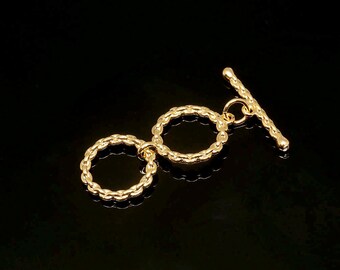 May include: A gold-coloured jewellery clasp with a textured chain-link design. The clasp features two circular rings connected to a bar with rounded ends. The background is black.