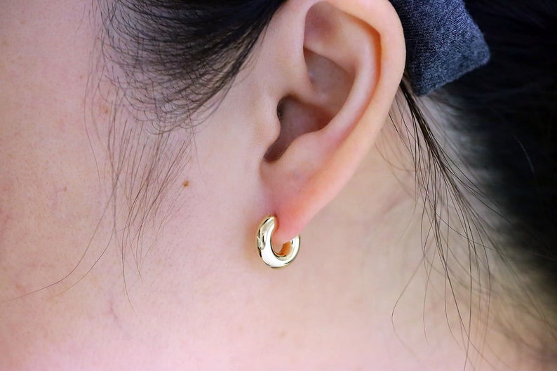 Puede incluir: Primer plano de una oreja con un pendiente de aro dorado. El pendiente tiene una forma peque&ntilde;a y redondeada, y est&aacute; bien sujeto a la oreja. La piel alrededor de la oreja es clara.