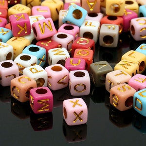 May include: Assortment of colorful alphabet letter beads, each bead is a cube with a gold letter on one side. The beads are in various colors, including pink, blue, yellow, green, and white.