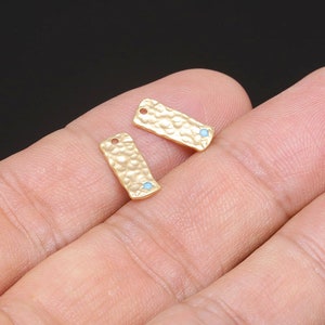 Op de afbeelding: Twee rechthoekige metalen labels in goudkleur met een gehamerde textuur en een klein turquoiseblauw steentje in het midden.