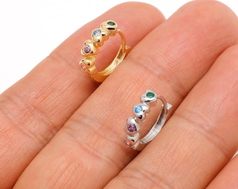 May include: Two small hoop earrings, one gold-toned and one silver-toned, each adorned with heart-shaped gemstones in various colors. The earrings are held in a hand, showcasing their design and details.