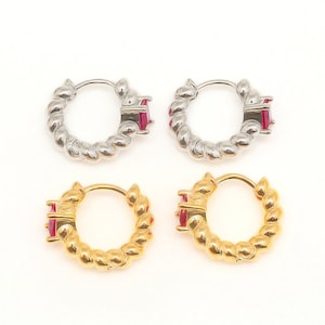 May include: Four hoop earrings with a twisted design. Two are silver with a red gemstone and two are gold with a red gemstone.