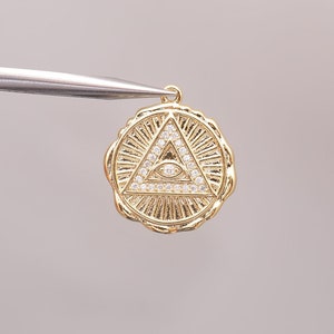Puede incluir: Colgante dorado con forma de triángulo con un diseño de ojo, rodeado de un círculo de diamantes de imitación transparentes.