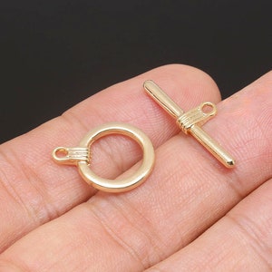 Puede incluir: Cierre de palanca de metal dorado con un anillo redondo y una barra recta con un bucle en un extremo. El cierre está diseñado para la fabricación de joyas.