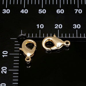 Puede incluir: Dos cierres de langosta de metal dorado, cada uno mide aproximadamente 2,5 cm de largo. Los cierres se muestran sobre un fondo negro con una regla para la escala.