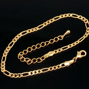 May include: Gold-colored anklet with a Figaro chain design. The anklet features a series of elongated and rounded links, with a lobster clasp closure and an adjustable chain. The jewelry is displayed against a black background.