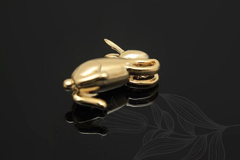 Puede incluir: Un amuleto de conejo de color dorado con la cola enroscada y una peque&ntilde;a oreja que sobresale. El amuleto est&aacute; tumbado de lado.