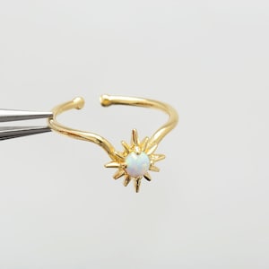 May include: A gold ring featuring a sunburst design with a central white gemstone. The ring is held by tweezers against a white backdrop. The ring's band is adjustable.