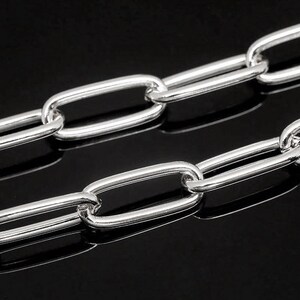 Puede incluir: Primer plano de una cadena plateada con eslabones ovalados alargados. La cadena es muy reflectante, con un acabado pulido. Los eslabones están conectados en un patrón repetitivo, creando un diseño elegante y moderno. El fondo es negro.