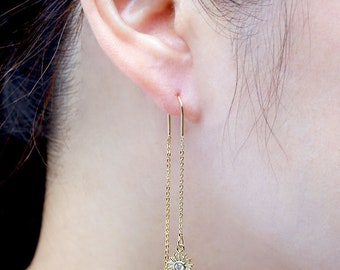 May include: Gold dangle earrings with a small sun charm featuring a clear crystal in the center. The sun charm has a textured, rayed design.