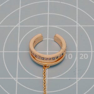 May include: A gold-toned ear cuff featuring a double-band design with a row of small, clear, sparkling stones embedded in the upper band. A delicate gold-toned chain hangs from the bottom of the cuff, which is positioned on a grid mat.