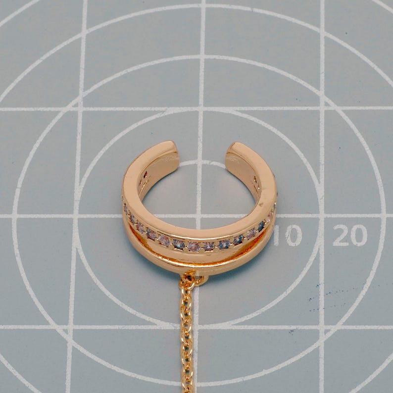 May include: A gold-toned ear cuff featuring a double-band design with a row of small, clear, sparkling stones embedded in the upper band. A delicate gold-toned chain hangs from the bottom of the cuff, which is positioned on a grid mat.