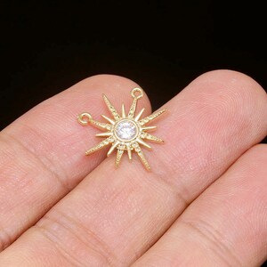 Könnte beinhalten: Ein goldfarbener Sonnenaufgangs-Anhänger mit einem zentralen, klaren Edelstein und spitzen Strahlen. Der Anhänger hat zwei kleine Schlaufen zum Befestigen an Schmuck. Der Anhänger wird in einer Hand vor schwarzem Hintergrund gehalten.