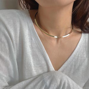 May include: A gold-colored, flat choker necklace. The necklace is a simple, elegant design, resting on a person's neck. The person is wearing a white, long-sleeved shirt with a V-neck.