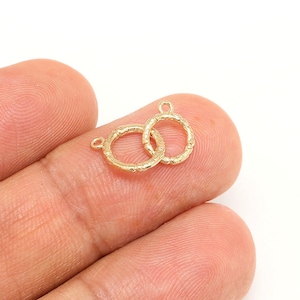 Könnte beinhalten: Zwei kleine, goldfarbene, offene Kreisanhänger mit strukturierten Details. Jeder Anhänger hat oben eine kleine Schlaufe zum Befestigen an Schmuck. Die Anhänger werden vor einem weißen Hintergrund gezeigt.