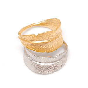 Puede incluir: Dos anillos en forma de pluma, uno dorado y otro plateado, apilados sobre un fondo blanco. Los anillos tienen una superficie texturizada, con el anillo dorado en la parte superior. Los anillos probablemente son de metal y están diseñados para ser usados como joyas.