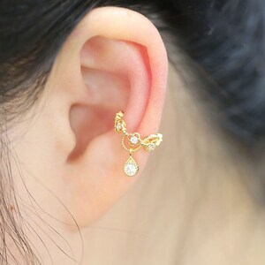 Peut inclure: Une manchette d'oreille dorée avec une goutte de cristal pendante. La manchette d'oreille est décorée de petits cristaux transparents.