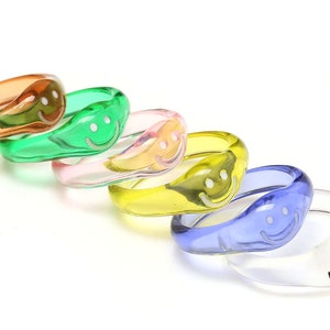 Pode incluir: Conjunto de seis anéis de plástico coloridos com carinhas sorridentes. Os anéis são em tons de laranja, verde, rosa, amarelo, azul e transparente.