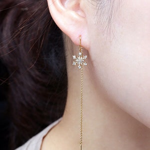 May include: A gold threader earring featuring a delicate snowflake charm adorned with small clear stones. The earring consists of a thin gold chain that passes through the earlobe, ending in a straight metal bar at the bottom of the chain.