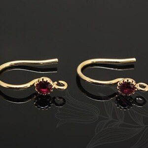 Pode incluir: Um par de ganchos de brinco de ouro com uma pequena gema vermelha em cada gancho.
