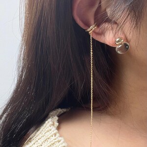 May include: A gold-toned ear cuff featuring a delicate, long dangling chain and a small, clear, round stud earring. The ear cuff is embellished with tiny, sparkling white stones along its band, creating a refined and layered appearance.
