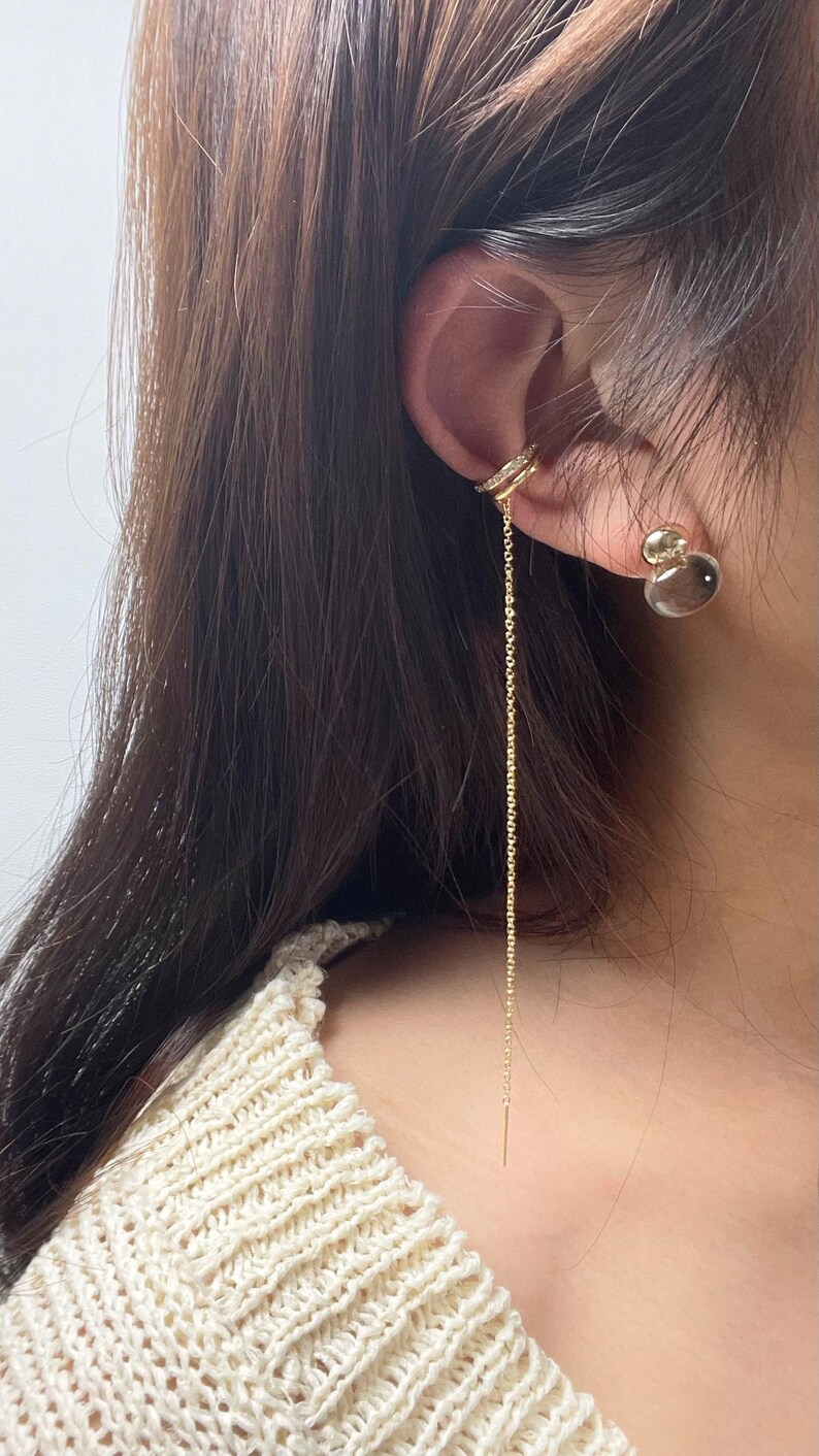 May include: A gold-toned ear cuff featuring a delicate, long dangling chain and a small, clear, round stud earring. The ear cuff is embellished with tiny, sparkling white stones along its band, creating a refined and layered appearance.
