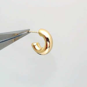 Puede incluir: Un solo pendiente de aro dorado brillante. El pendiente est&aacute; sujeto por unos alicates plateados sobre un fondo blanco. El pendiente tiene un dise&ntilde;o tubular redondeado.