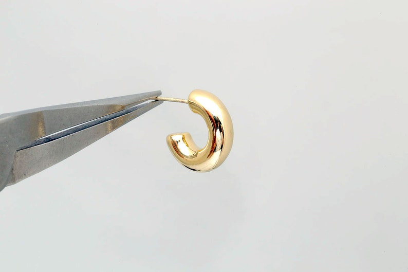 Puede incluir: Un solo pendiente de aro dorado brillante. El pendiente est&aacute; sujeto por unos alicates plateados sobre un fondo blanco. El pendiente tiene un dise&ntilde;o tubular redondeado.