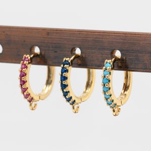 May include: Three gold hoop earrings with a row of colorful gemstones. The earrings are displayed on a wooden stand. The earrings are available in pink, blue, and turquoise.