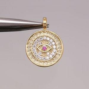 May include: Gold pendant charm with a round, ornate design featuring a white and pink evil eye with a single pink stone in the center.