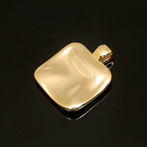 May include: A gold-colored pendant with a square, slightly curved shape. The pendant has a loop at the top for a chain. The surface is smooth and reflective, with a polished finish. The background is black.