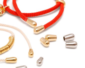 May include: A bright red cord and a cream-colored cord, both featuring polished gold-tone metal end caps. Several loose, bullet-shaped metal cord ends in gold and silver tones are scattered nearby, showcasing their smooth, rounded, and hollow design.