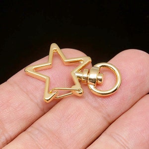 Puede incluir: Un cierre giratorio en forma de estrella de color dorado, con un anillo circular y un cierre de bisagra. La estrella tiene cinco puntas y un acabado liso y pulido. El cierre está diseñado para sujetarse a una variedad de artículos.