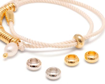 May include: A beige rope bracelet featuring a series of gold-toned cylindrical beads and a single white pearl. The bracelet includes a sliding gold-toned bead for adjustment, accompanied by four loose, polished metal beads in gold and silver tones.