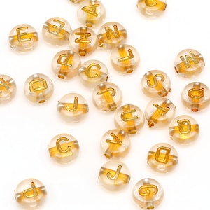 May include: A collection of translucent round beads with gold letters spelling out the alphabet. The beads are scattered on a white surface.