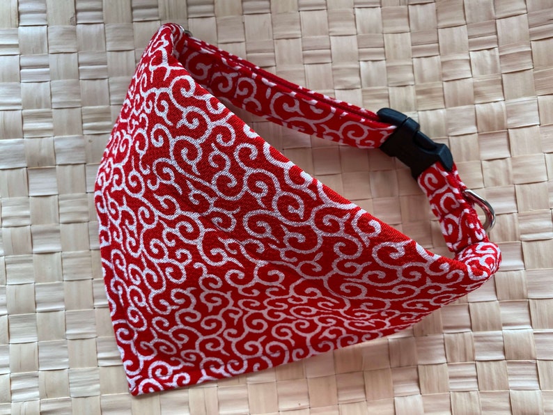 Puede incluir: Collar para mascotas con estampado rojo y blanco con hebilla de pl&aacute;stico negro y anilla met&aacute;lica plateada. El collar presenta un dise&ntilde;o de remolino repetido. El fondo es una superficie tejida de color marr&oacute;n claro.