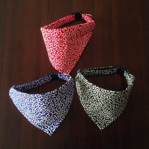 Puede incluir: Tres pa&ntilde;uelos para perros en rojo, azul y negro, cada uno con un patr&oacute;n de remolino blanco. Los pa&ntilde;uelos son triangulares y tienen collares negros ajustables. El pa&ntilde;uelo rojo est&aacute; en la parte superior, con los pa&ntilde;uelos azul y negro debajo.