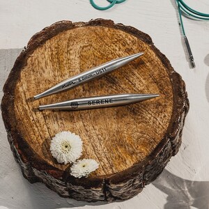 May include: Two silver knitting needles on a wooden surface. The larger needle is labelled "KnitPro 8.0mm - US11". The smaller needle is labelled "SERENE".