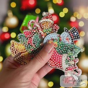 May include: A clear Christmas ornament featuring a Santa Claus, snowman, and reindeer design. The ornament is decorated with red, green, and white rhinestones. The ornament is held in a hand in front of a blurred Christmas tree.