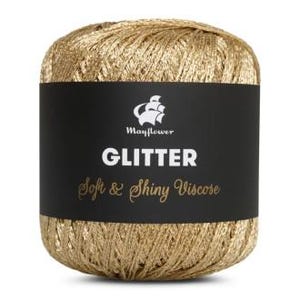 May include: A ball of gold-colored yarn with a black label wrapped around it. The label reads "Mayflower GLITTER Soft & Shiny Viscose" in white text. The yarn appears soft and has a shiny, metallic appearance.