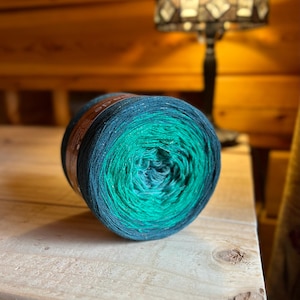 May include: A skein of yarn in a gradient of teal, blue, and green colors. The yarn is wound into a ball shape and is ready for knitting or crocheting.