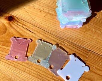 Könnte beinhalten: Fünf Plastik-Garnrollen in verschiedenen Pastellfarben. Die Spulen sind durchscheinend und haben ein Loch in der Mitte.