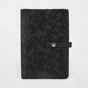 May include: A black, rectangular case with a floral pattern. The case has a strap with a silver snap closure. The floral design is a darker shade of black, creating a subtle contrast. The case is closed.