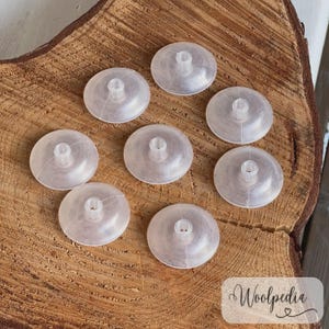 May include: A collection of eight clear, plastic yarn spool holders. Each holder is circular with a central post. The holders are arranged on a textured, wooden surface. The image includes the text "Woolpedia".