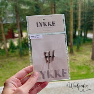 Peut inclure: Un emballage plastique transparent contenant des accessoires de tricotage de la marque LYKKE. L'emballage présente le logo LYKKE et les mots "MAKE HAPPY" et "ACCESSORIES". Il contient trois outils de tricotage en métal argenté. Le texte "Access Connect Cord" est également visible.