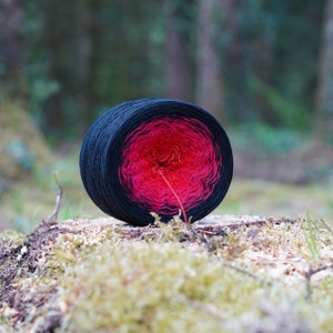 May include: A ball of yarn with a gradient color scheme, transitioning from black to red. The yarn is resting on a mossy tree stump in a natural outdoor setting. The yarn is likely used for knitting or crocheting.