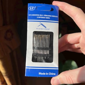Könnte beinhalten: Eine Packung mit 6/12 sortierten, selbst-einfädelnden Nadeln in drei verschiedenen Größen. Die Verpackung ist blau und weiß mit dem Text "JIN LI" und "So einfach ist das Einfädeln!" Die Nadeln befinden sich in einem schwarzen Plastiktablett. Die Verpackung trägt den Hinweis "Made in China".